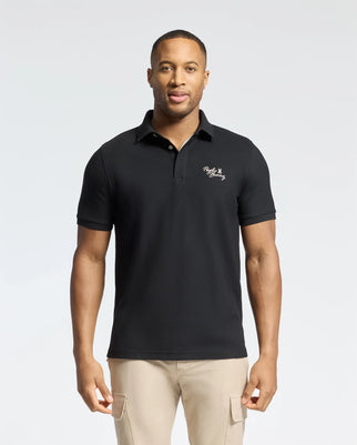 A man wears a Psycho Bunny MENS DESERT PIQUE POLO - B6K506F200, black with white chest embroidery, made from 100% Pima cotton, and pairs it with beige pants. The plain white background accentuates the timeless style.