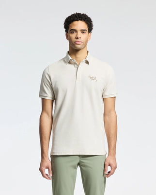A person stands in a Psycho Bunny light beige MENS DESERT PIQUE POLO - B6K506F200 made from luxurious Pima cotton with an embroidered chest logo, paired with olive green pants. The plain white background accentuates the timeless style of the ensemble.