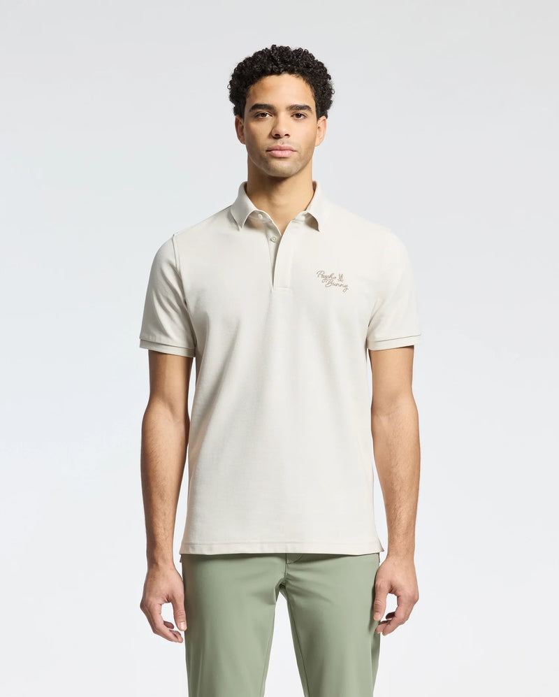 A person stands in a Psycho Bunny light beige MENS DESERT PIQUE POLO - B6K506F200 made from luxurious Pima cotton with an embroidered chest logo, paired with olive green pants. The plain white background accentuates the timeless style of the ensemble.