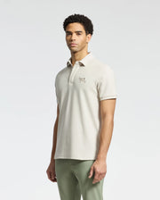 Someone wears a Psycho Bunny MENS DESERT PIQUE POLO in light beige, crafted from luxurious Pima cotton with an embroidered chest logo, paired seamlessly with light green pants. The timeless style stands out against the plain white backdrop.