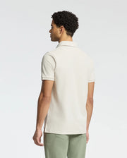 A man wearing a Psycho Bunny MENS DESERT PIQUE POLO - B6K506F200, made of Pima cotton in a classic design, stands with his back to the camera against a plain white background.
