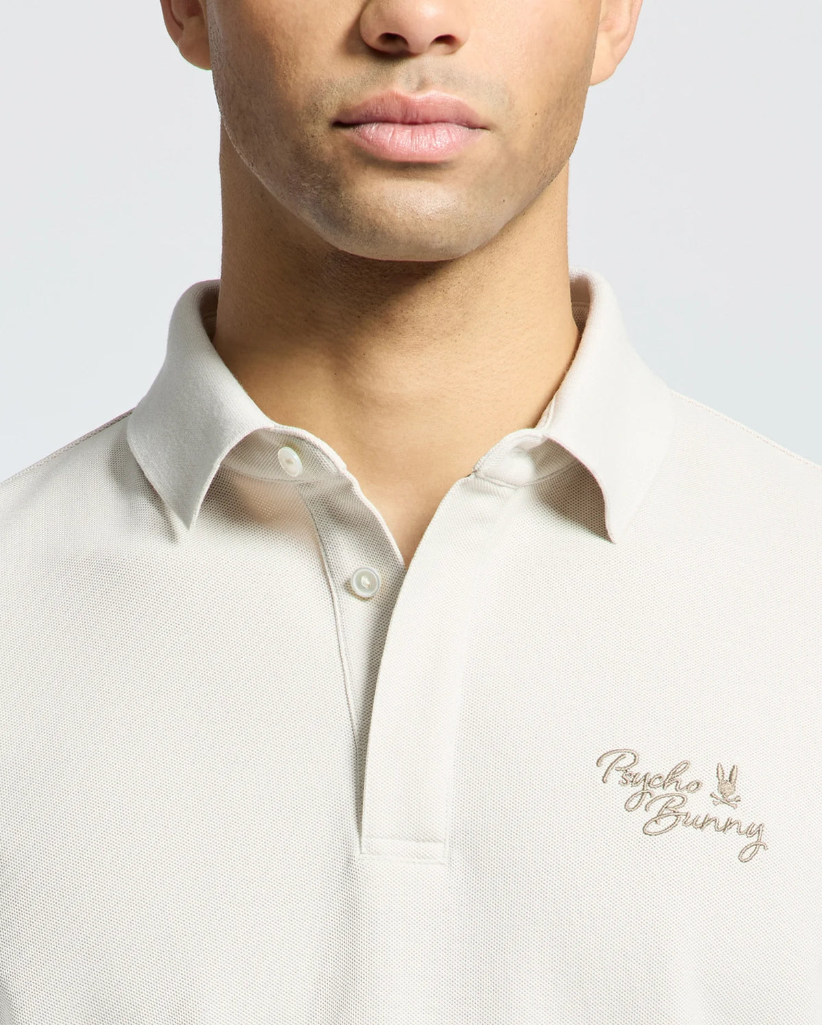 A man wears Psycho Bunny's MENS DESERT PIQUE POLO (B6K506F200) in beige, featuring embroidered text and logo on the chest, highlighting its classic style against a light gray backdrop.