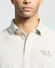 A man wears Psycho Bunny's MENS DESERT PIQUE POLO (B6K506F200) in beige, featuring embroidered text and logo on the chest, highlighting its classic style against a light gray backdrop.