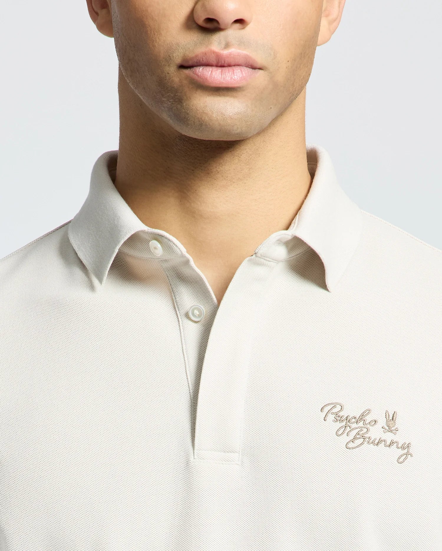 A man wears Psycho Bunny's MENS DESERT PIQUE POLO (B6K506F200) in beige, featuring embroidered text and logo on the chest, highlighting its classic style against a light gray backdrop.