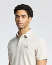 A person with short, curly hair wears Psycho Bunny's MENS DESERT PIQUE POLO - B6K506F200 in beige Pima cotton, featuring a small embroidered logo on the chest. The neutral backdrop reflects their timeless style and calm expression.