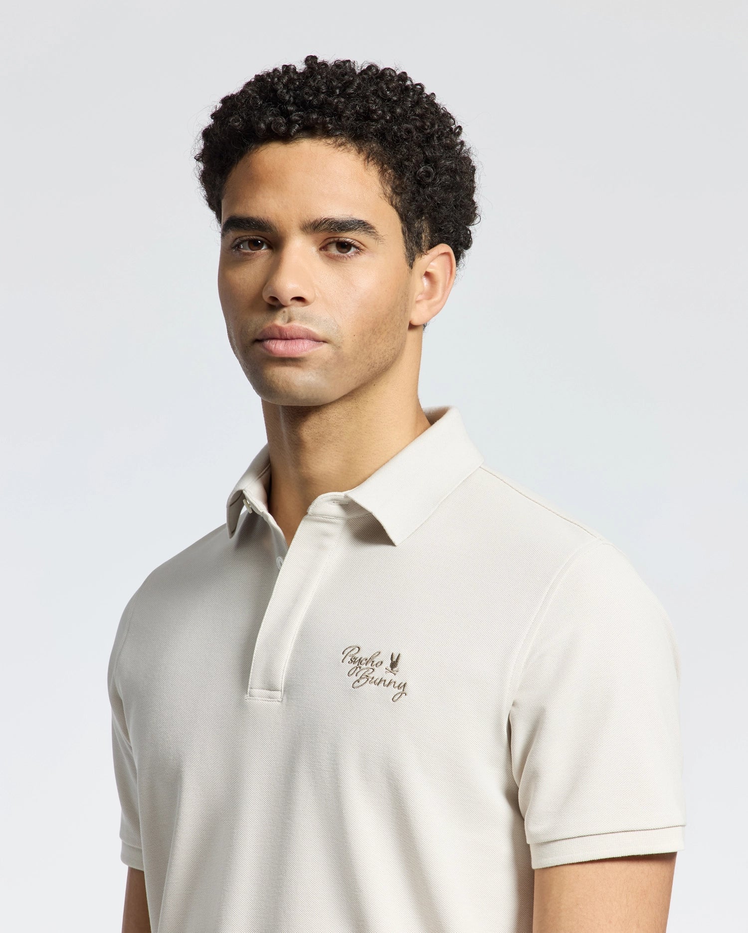 A person with short, curly hair wears Psycho Bunny's MENS DESERT PIQUE POLO - B6K506F200 in beige Pima cotton, featuring a small embroidered logo on the chest. The neutral backdrop reflects their timeless style and calm expression.