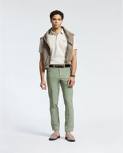A man stands showcasing timeless style in a Psycho Bunny MENS DESERT PIQUE POLO - B6K506F200 made from soft Pima cotton, with a taupe jacket draped over his shoulders. He wears light green pants, a brown belt, and light brown loafers against a plain white background.