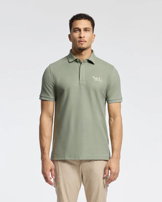 A man in a sage green Psycho Bunny MENS DESERT PIQUE POLO - B6K506F200, featuring a subtle chest logo, stands against a light background. With short curly hair and beige pants, he effortlessly embodies timeless style.