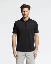 A man with short brown hair and a beard wears the Psycho Bunny MENS TREVOR PERFORMANCE PIQUE ZIP UP POLO (B6K557F200) in black and light gray pants, standing against a plain light background, facing the camera with a neutral expression.