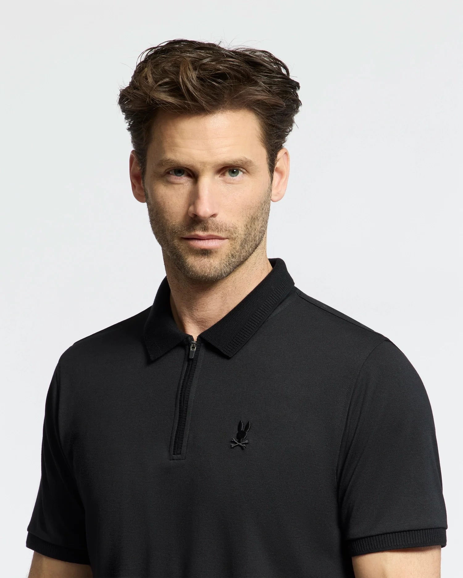A man with short brown hair and light stubble wears the Psycho Bunny MENS TREVOR PERFORMANCE PIQUE ZIP UP POLO (B6K557F200), a black slim-fit collared shirt with an embroidered bird logo, standing against a plain light background.