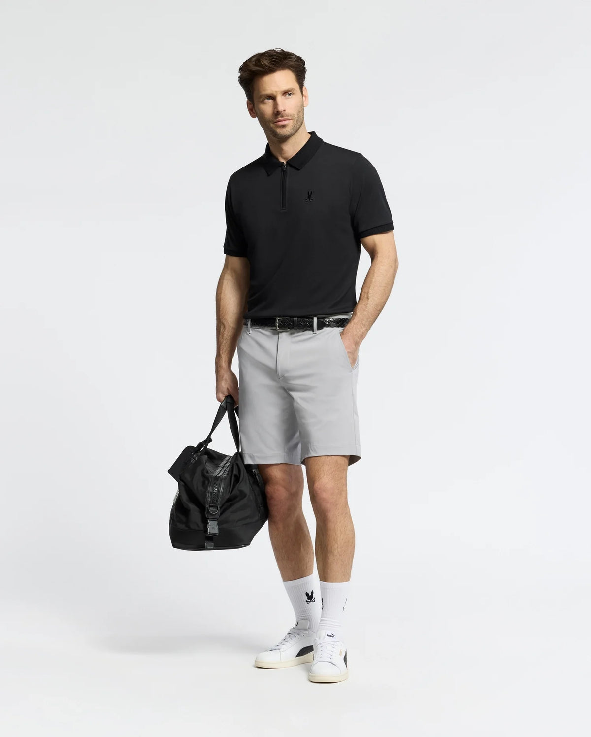 A man stands against a plain background wearing the Psycho Bunny MENS TREVOR PERFORMANCE PIQUE ZIP UP POLO in black, light gray shorts, white socks with a black logo, white sneakers, and holding a black duffel bag by his side.
