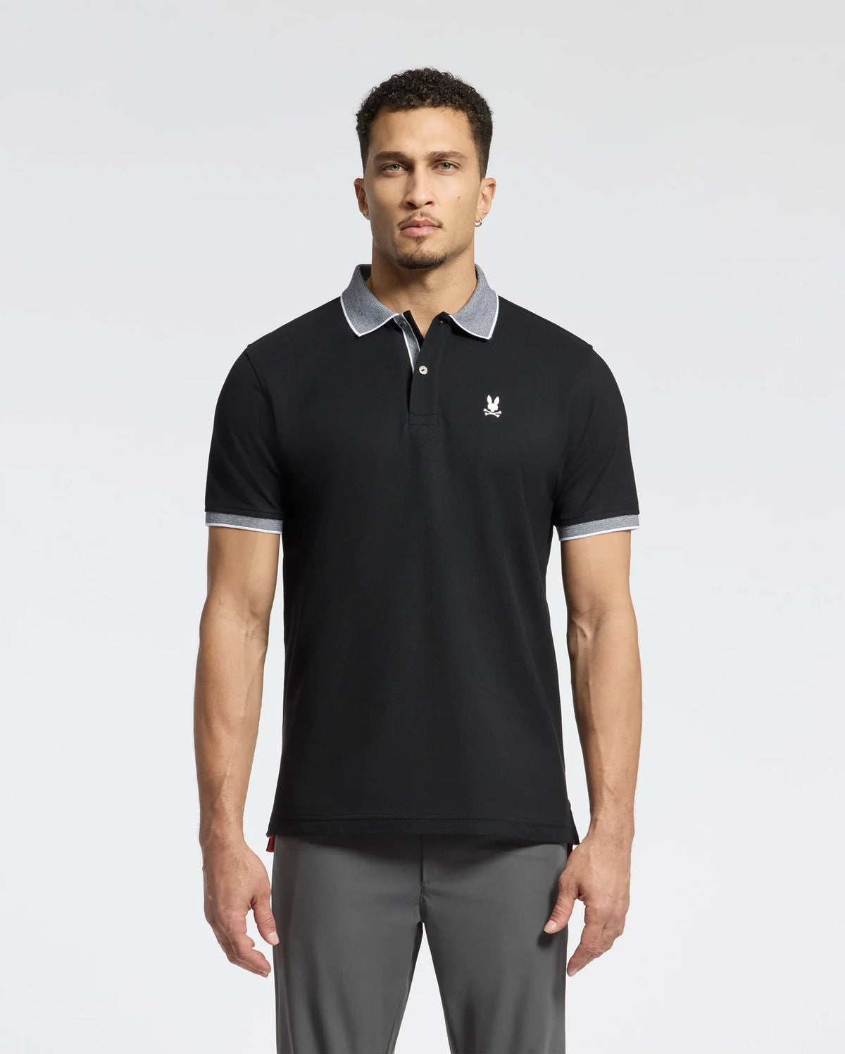 A man wears a Psycho Bunny MENS JONAS STRIPE COLLAR PIQUE POLO - B6K583F200. It's black, made from luxurious Pima Cotton with a gray collar and sleeve trim, featuring a small white emblem on the chest, showcasing its premium Peruvian quality against a plain white backdrop.