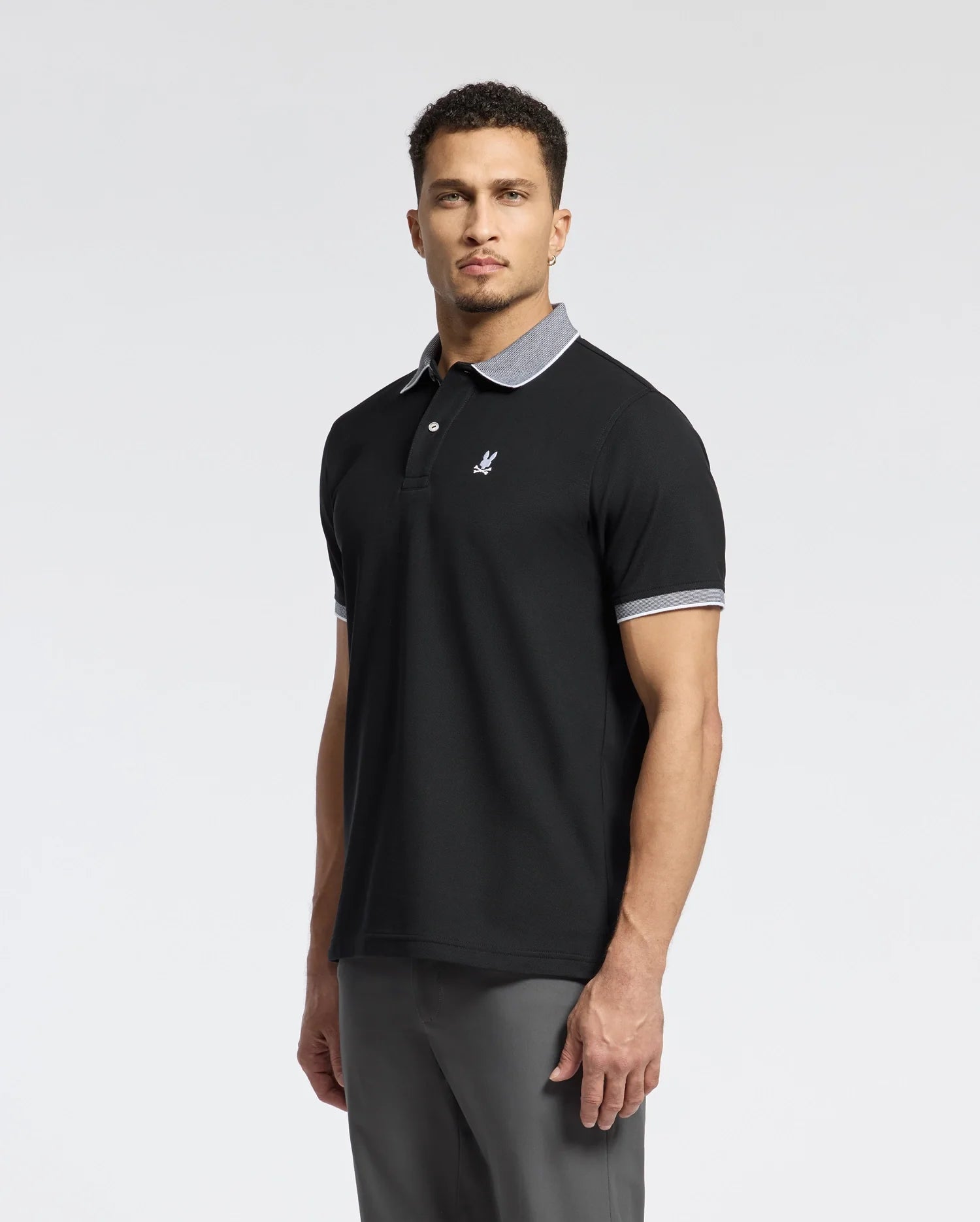 A man with short curly hair models the Psycho Bunny Men's Jonas Stripe Collar Pique Polo (B6K583F200), crafted from luxurious Peruvian Pima Cotton. It features a black design, gray collar and cuffs with micro-stripe detail, and an embroidered chest logo. He stands against a plain white background, looking at the camera.