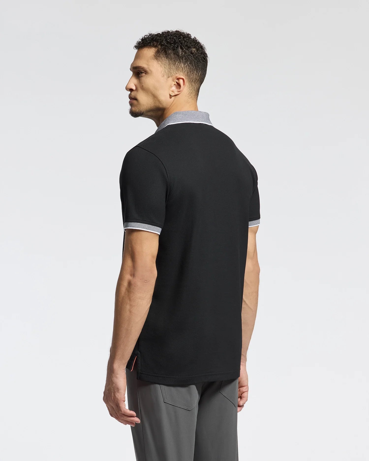 A man with short curly hair stands gazing sideways, wearing the Psycho Bunny MENS JONAS STRIPE COLLAR PIQUE POLO in black with grey trim, crafted from soft Pima cotton. His gray pants complement the look against a white background, subtly hinting at Peruvian elegance with their micro-stripe detail.