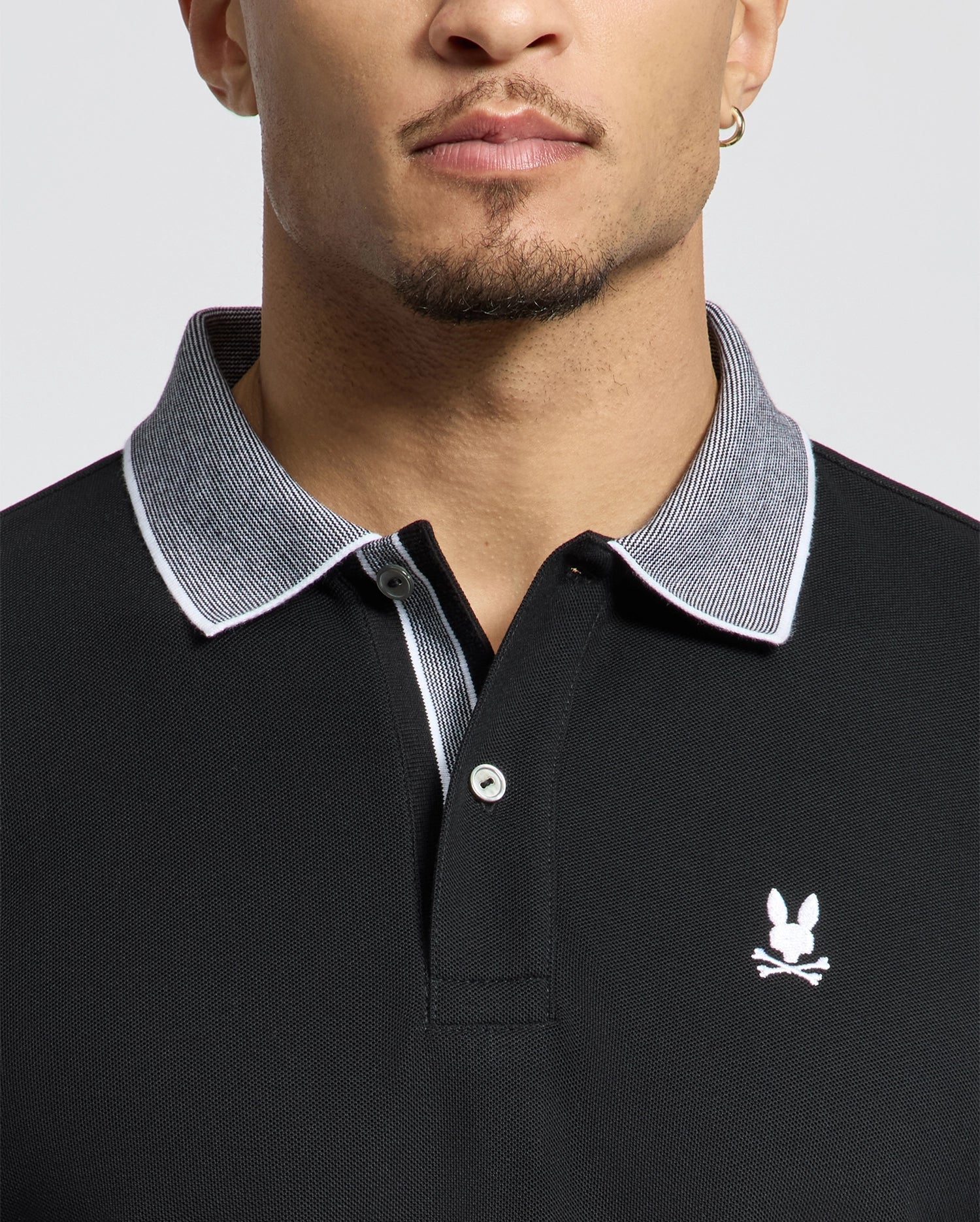 A person wearing a black Psycho Bunny MENS JONAS STRIPE COLLAR PIQUE POLO with a gray collar and small white bunny logo above crossed bones. Made from Pima Cotton, it features micro-stripes. The focus is on the upper torso and neck, showcasing short hair and a stud earring.
