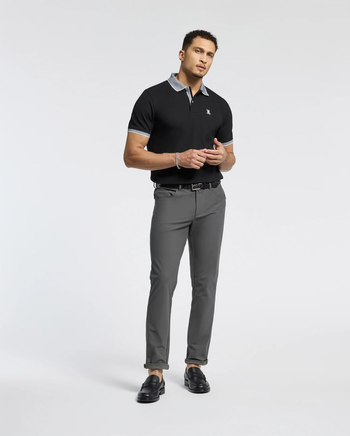 A man stands against a plain background wearing a Psycho Bunny Mens Jonas Stripe Collar Pique Polo in black, featuring a gray collar and cuffs. He pairs it with gray pants and black loafers, holding an item near his waist.