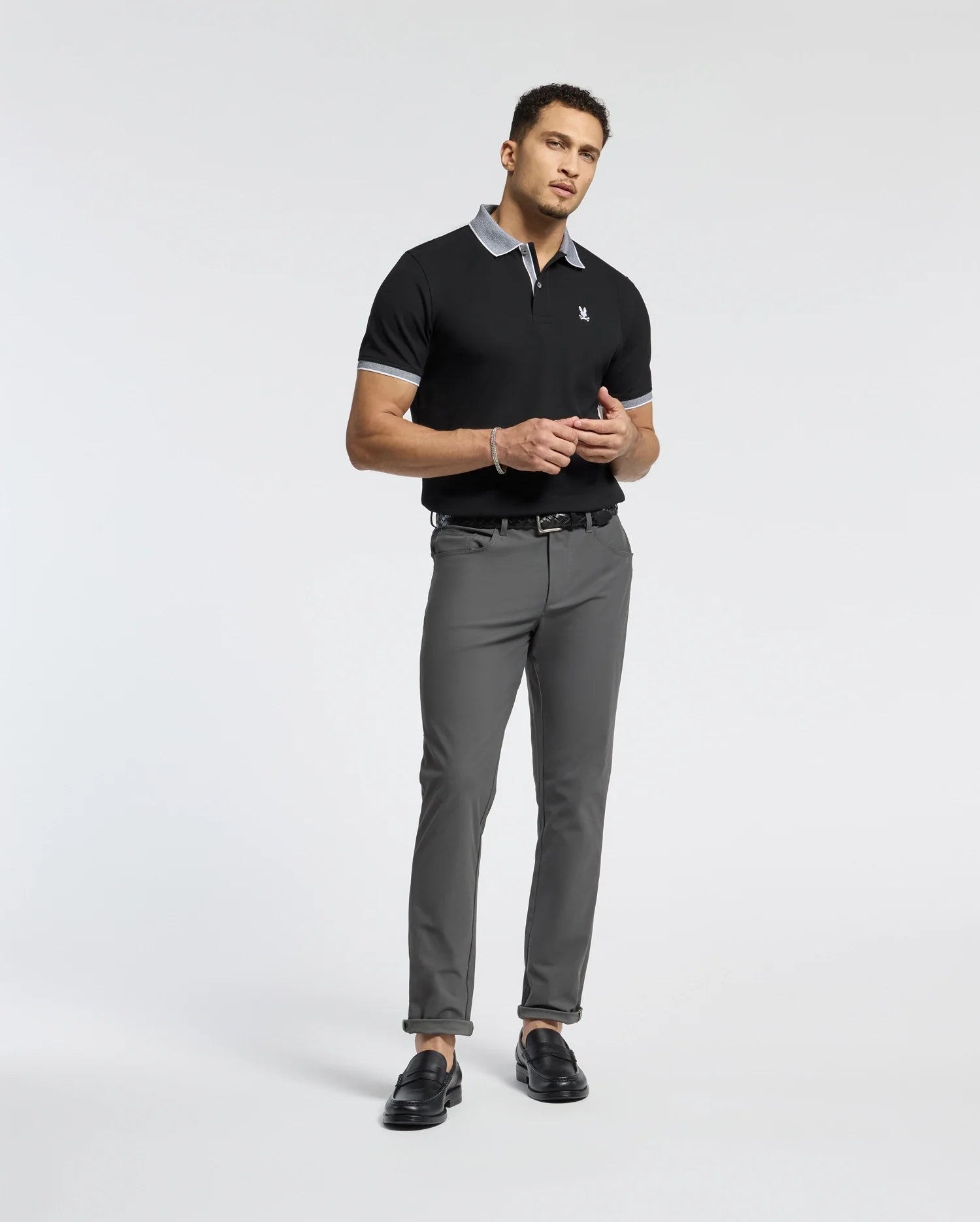 A man stands against a plain background wearing a Psycho Bunny Mens Jonas Stripe Collar Pique Polo in black, featuring a gray collar and cuffs. He pairs it with gray pants and black loafers, holding an item near his waist.