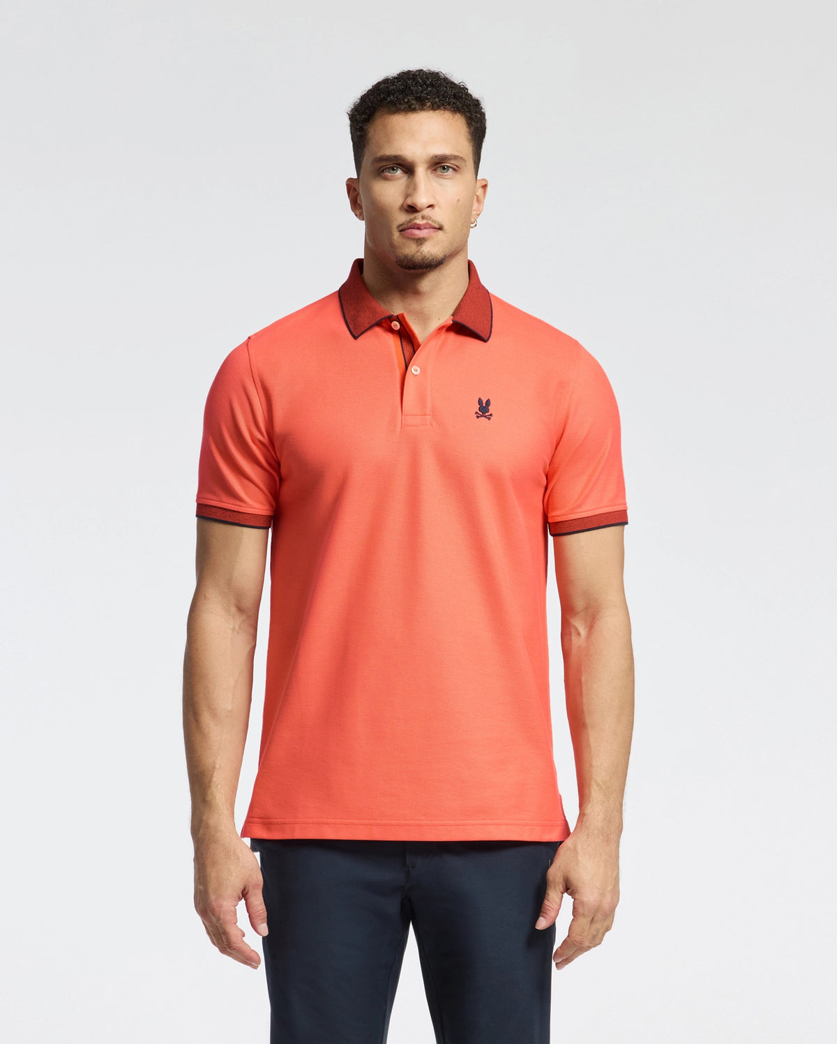 A man models a Psycho Bunny MENS JONAS MICRO STRIPE COLLAR PIQUE POLO in orange, made from Pima cotton with dark collar and sleeve trim. It features a micro-stripe pattern, regular fit, small logo detail, and is paired with dark pants against a plain background.