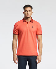 A man models a Psycho Bunny MENS JONAS MICRO STRIPE COLLAR PIQUE POLO in orange, made from Pima cotton with dark collar and sleeve trim. It features a micro-stripe pattern, regular fit, small logo detail, and is paired with dark pants against a plain background.