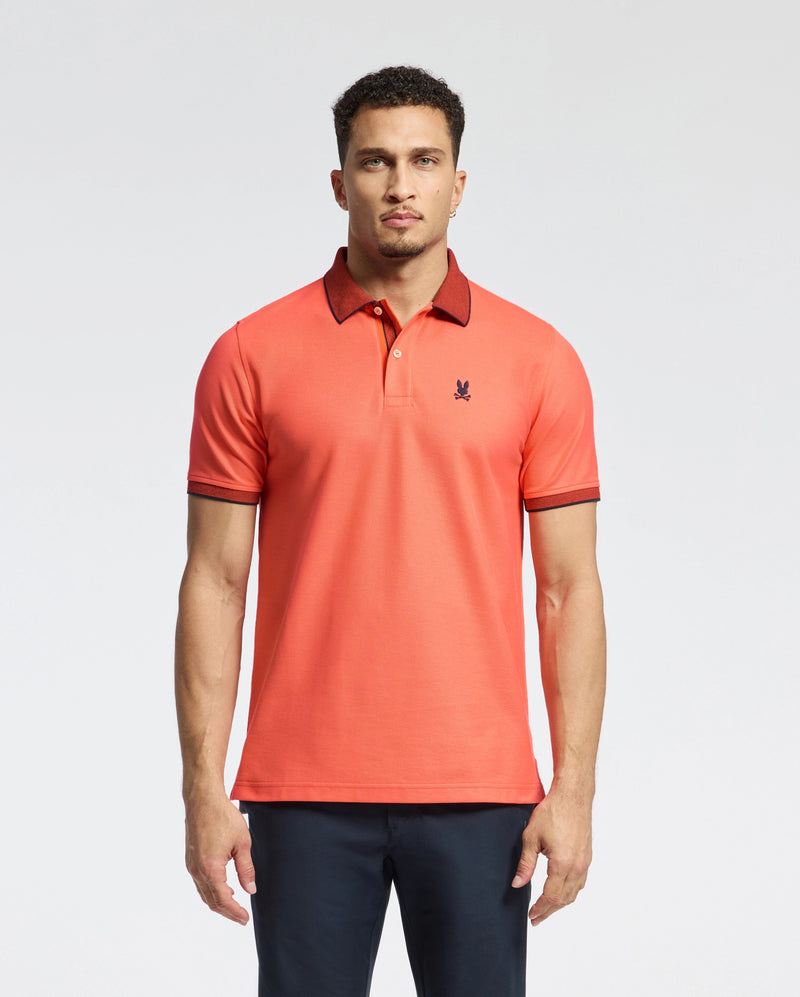 A man models a Psycho Bunny MENS JONAS MICRO STRIPE COLLAR PIQUE POLO in orange, made from Pima cotton with dark collar and sleeve trim. It features a micro-stripe pattern, regular fit, small logo detail, and is paired with dark pants against a plain background.
