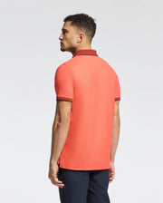 A person is wearing the Psycho Bunny MENS JONAS MICRO STRIPE COLLAR PIQUE POLO B6K583F200, a regular fit short-sleeved coral Pima cotton shirt. It features dark micro-striped collar and sleeve edges. The person stands facing slightly right on a plain light background with arms relaxed.