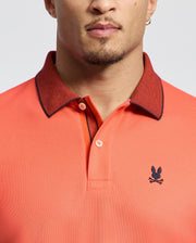 A person wearing the Psycho Bunny MENS JONAS MICRO STRIPE COLLAR PIQUE POLO in coral, featuring micro-stripes, a dark collar, and a small stitched rabbit and crossbones emblem. The partially visible face adds mystery to the casual vibe.