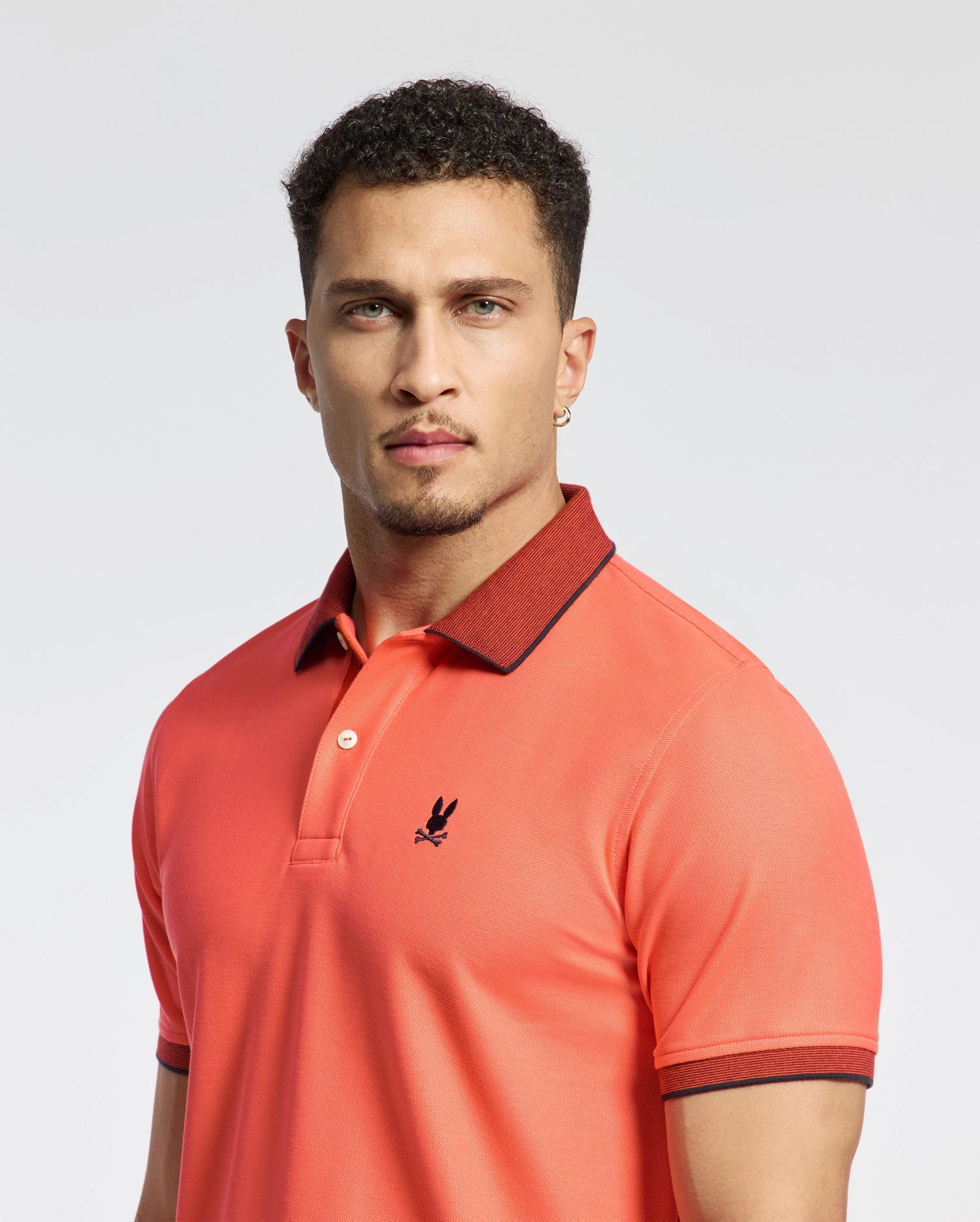 A man in a Psycho Bunny Jonas polo made from Pima cotton with a micro stripe collar and embroidered logo stands against a light background. The regular fit accents his short curly hair, and he's wearing a small hoop earring.