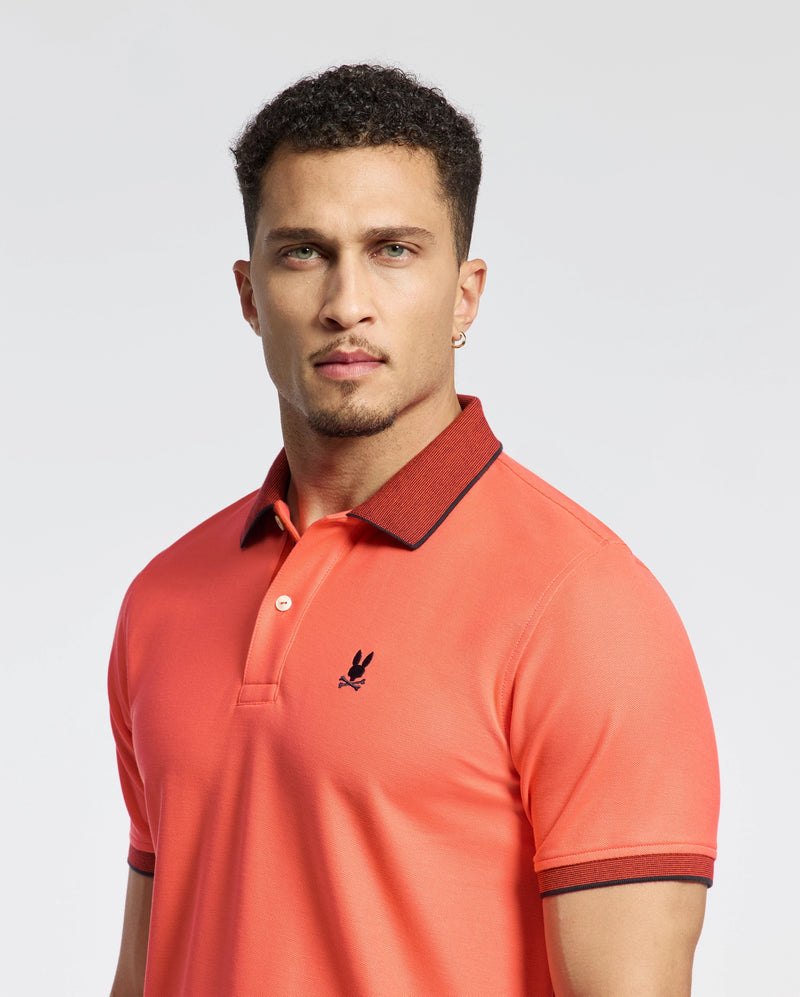 A man in a Psycho Bunny Jonas polo made from Pima cotton with a micro stripe collar and embroidered logo stands against a light background. The regular fit accents his short curly hair, and he's wearing a small hoop earring.