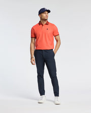 A man in a Psycho Bunny MENS JONAS MICRO STRIPE COLLAR PIQUE POLO - B6K583F200 stands against a plain background, wearing navy pants, a navy cap, and white sneakers. His left hand is in his pocket as he gazes right. The regular fit polo's subtle micro-stripe detailing enhances the look.