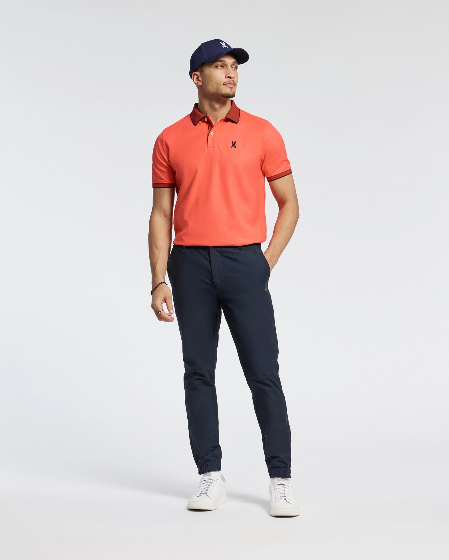 A man in a Psycho Bunny MENS JONAS MICRO STRIPE COLLAR PIQUE POLO - B6K583F200 stands against a plain background, wearing navy pants, a navy cap, and white sneakers. His left hand is in his pocket as he gazes right. The regular fit polo's subtle micro-stripe detailing enhances the look.