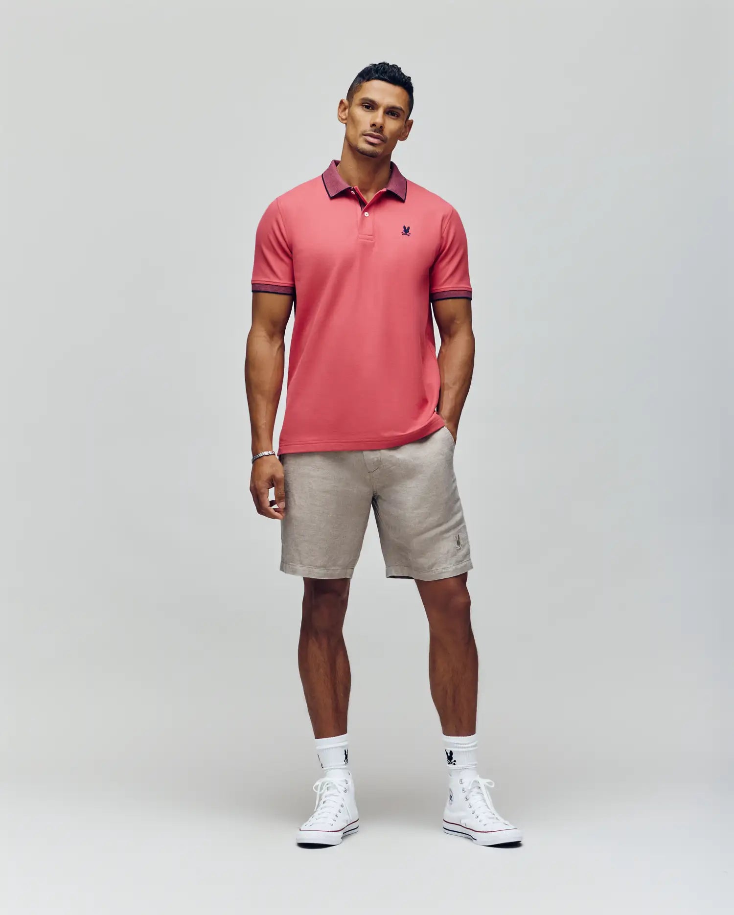 A man with short hair and a neutral expression stands against a plain background wearing the Psycho Bunny MENS JONAS MICRO STRIPE COLLAR PIQUE POLO in pink, paired with beige shorts, white socks, white sneakers, and one hand in his pocket.