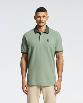A man in a sage green Psycho Bunny MENS JONAS MICRO STRIPE COLLAR PIQUE POLO, featuring a dark collar, cuffs, and an embroidered chest logo, stands against a plain backdrop. He pairs the Peruvian Pima cotton shirt with beige pants and maintains a neutral expression.