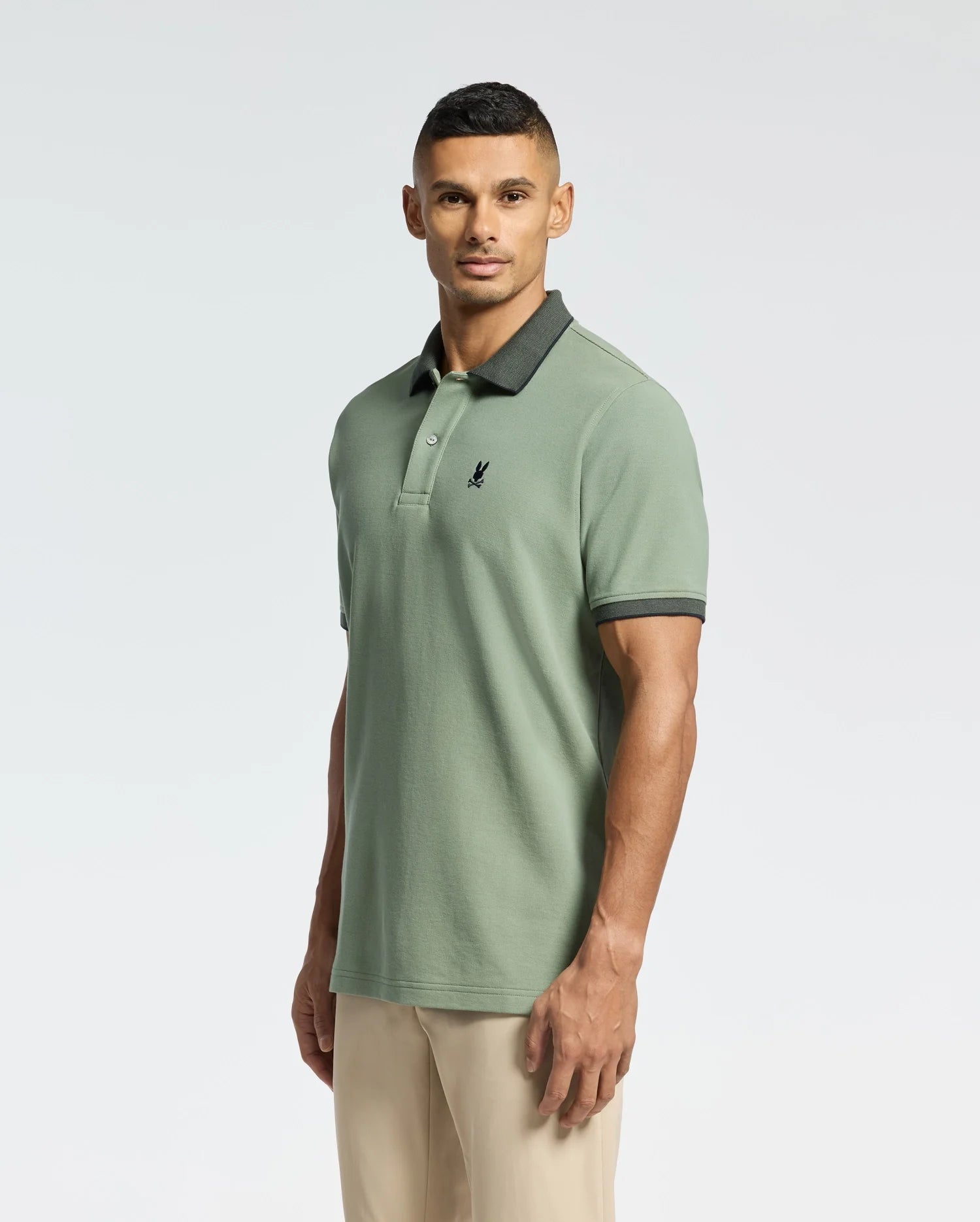 Against a plain backdrop, a person in Psycho Bunny's MENS JONAS MICRO STRIPE COLLAR PIQUE POLO (B6K583F200) made from Peruvian Pima cotton stands wearing beige pants. The light green polo features a dark collar and emblem. They have short hair and gaze directly at the camera.