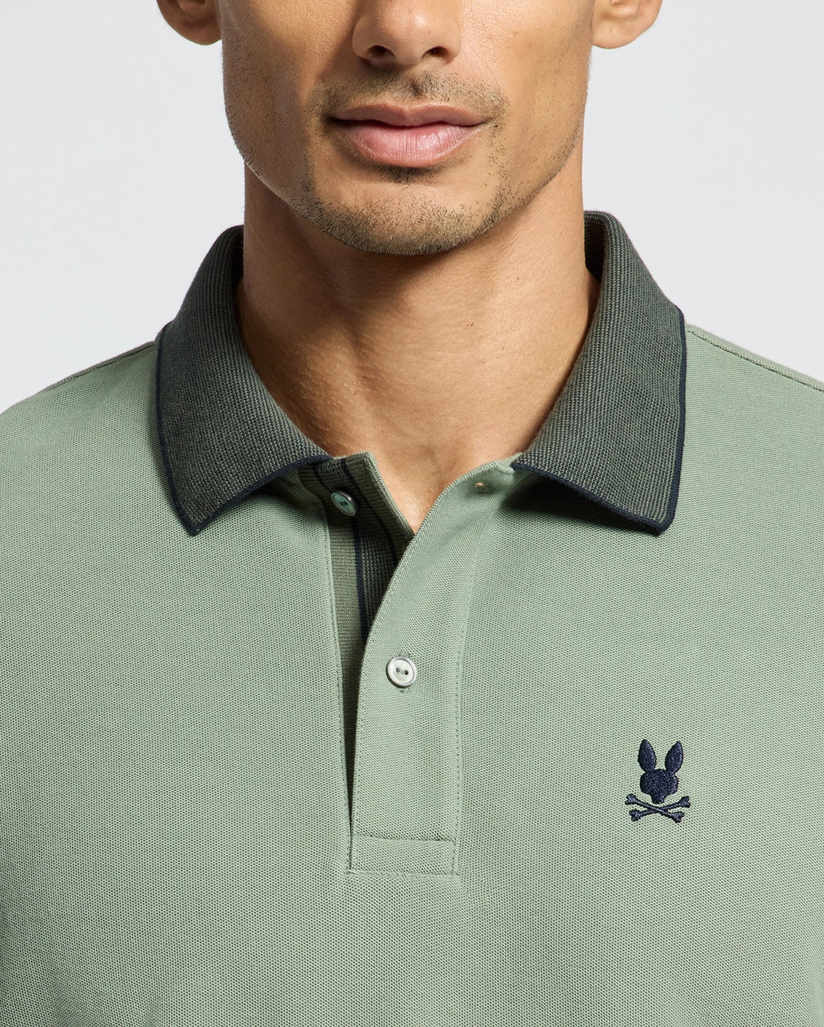 A man wears a green Psycho Bunny B6K583F200 polo with a dark collar, made from soft Pima Cotton. It features a small embroidered bunny logo above crossed bones on the left. The focus is on the upper torso, excluding the full face.