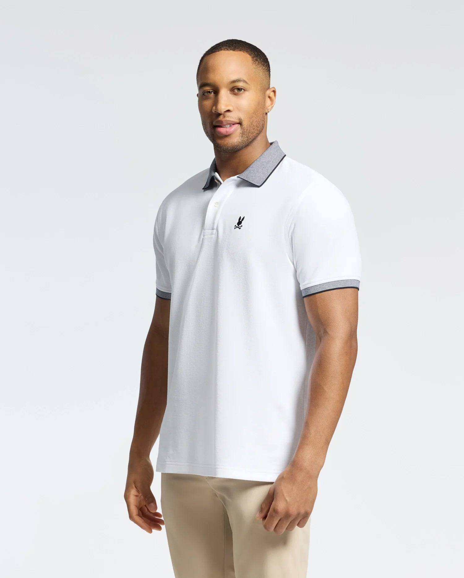 A man stands against a light background wearing the Psycho Bunny MENS JONAS STRIPE COLLAR PIQUE POLO (B6K583F200) with dark trim on the collar and sleeves, beige pants, and a small chest logo.