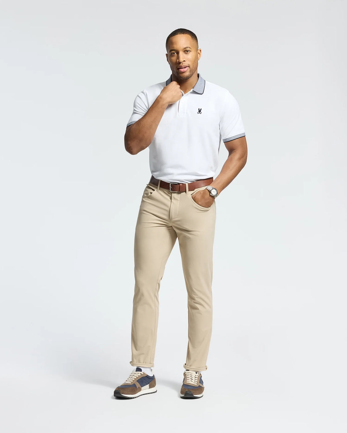 A man poses against a plain background wearing the Psycho Bunny MENS JONAS STRIPE COLLAR PIQUE POLO (B6K583F200), beige pants, a brown belt, watch, and sneakers. He rests one hand on his neck and looks at the camera.