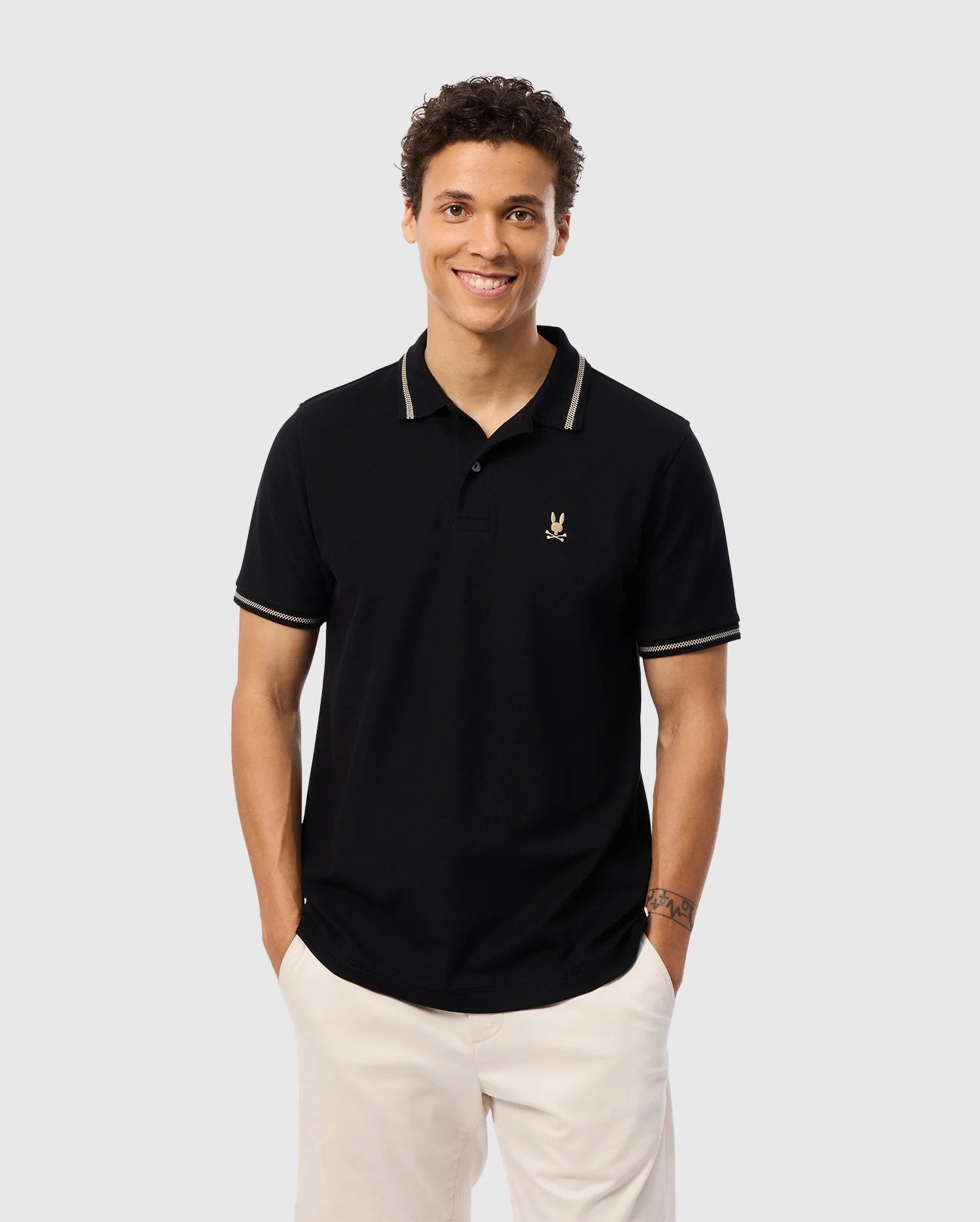 A smiling person wears a Psycho Bunny Men's Riviera Pique Polo Shirt made from soft Pima cotton, featuring an embroidered Bunny logo on the chest. The shirt includes light-colored collar trim and memory cuffs, styled with light trousers against a plain gray background.