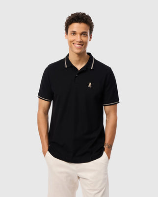 A smiling person wears a Psycho Bunny Men's Riviera Pique Polo Shirt made from soft Pima cotton, featuring an embroidered Bunny logo on the chest. The shirt includes light-colored collar trim and memory cuffs, styled with light trousers against a plain gray background.