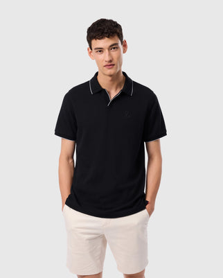 Standing with hands in pockets, a person wears the stylish Psycho Bunny Richards Johnny Collar Pique Polo Shirt in black with white trim, paired with beige shorts, against a simple gray background.