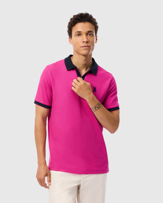 A man with short curly hair is wearing a vibrant pink Psycho Bunny Men's Richards Johnny Collar Pique Polo Shirt (B6K641C200) made of Pima cotton, featuring black trim on the collar and sleeves. He has a minimal wrist tattoo and stands against a plain light gray background. The regular fit shirt features a small black logo on the left chest.