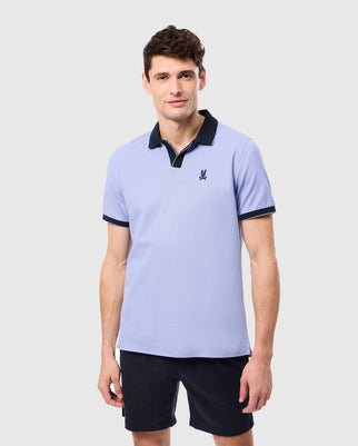 A person is wearing a Psycho Bunny regular fit light purple MENS RICHARDS JOHNNY COLLAR PIQUE POLO SHIRT, crafted from soft Pima cotton, featuring a dark collar and small chest logo, paired with dark shorts against a plain background.