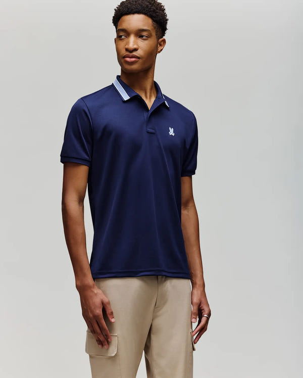 A man in a Psycho Bunny MENS FORREST SPORT PIQUE POLO (B6K660F200), navy blue with a white chest logo, and beige cargo pants stands against a light gray background, looking slightly left with a neutral expression.