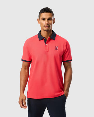 A man wearing a vibrant red Psycho Bunny Mens Kayden Pique Polo (B6K675C200) with a navy blue collar and sleeve edges. The shirt, made from luxurious Pima cotton piqué, features a small embroidered logo on the left chest. He has short hair and is standing with his hands in his pockets against a plain light gray background.