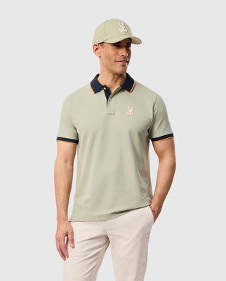 Man wearing a light green MENS KAYDEN PIQUE POLO - B6K675C200 by Psycho Bunny with a small embroidered bunny on the left chest, a matching light green baseball cap with the same logo, and beige pants. He is smiling and standing against a plain white background.