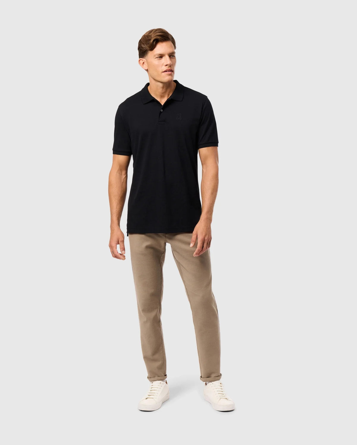 A man stands against a plain white background, wearing the Psycho Bunny MENS JAMES PREMIUM PIQUE POLO SHIRT - B6K945D200 in black, beige pants, and white sneakers. He is looking slightly to his left with a neutral expression and has short, neatly styled hair. His right hand rests by his side, and his left hand is slightly forward.