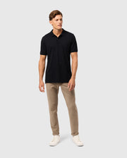 A man stands against a plain white background, wearing the Psycho Bunny MENS JAMES PREMIUM PIQUE POLO SHIRT - B6K945D200 in black, beige pants, and white sneakers. He is looking slightly to his left with a neutral expression and has short, neatly styled hair. His right hand rests by his side, and his left hand is slightly forward.