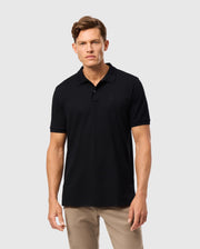 A man with short hair stands against a plain background, wearing the Psycho Bunny MENS JAMES PREMIUM PIQUE POLO SHIRT - B6K945D200 in black and beige pants. He faces the camera with a neutral expression.
