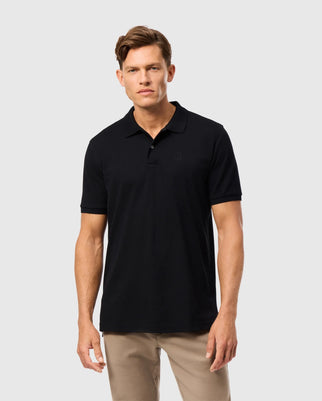 A man with short hair stands against a plain background, wearing the Psycho Bunny MENS JAMES PREMIUM PIQUE POLO SHIRT - B6K945D200 in black and beige pants. He faces the camera with a neutral expression.