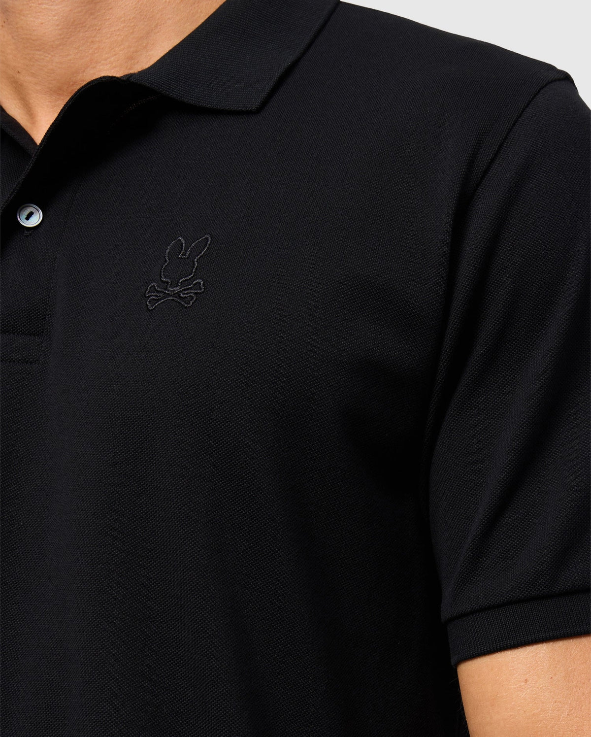 Close-up of a person wearing the Psycho Bunny MENS JAMES PREMIUM PIQUE POLO SHIRT - B6K945D200, crafted from durable Pima cotton in black. The shirt features a small, subtle embroidered bunny with crossbones logo on the left chest area. The background is plain and light-colored, showing only part of the chest and shoulder.