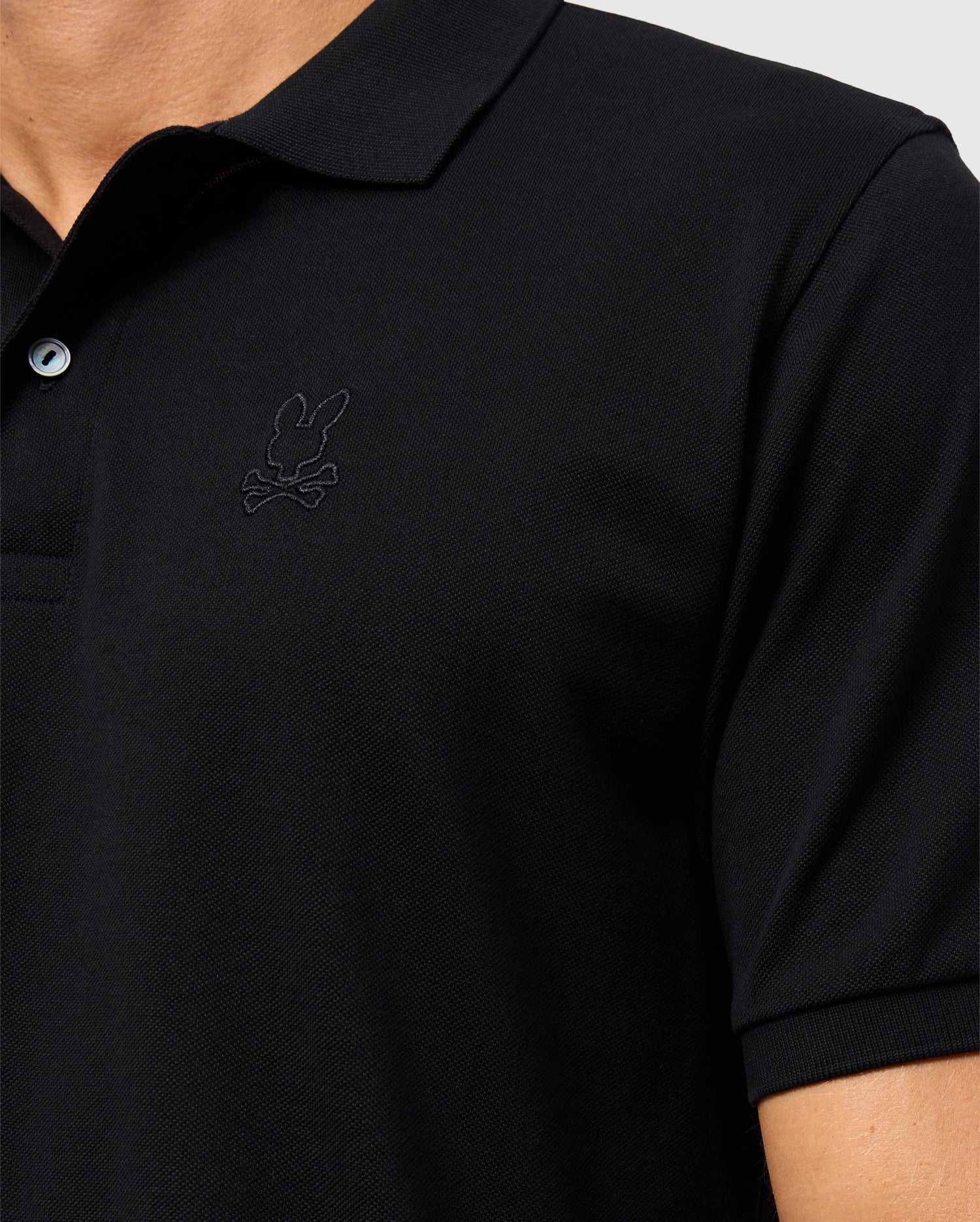 Close-up of a person wearing the Psycho Bunny MENS JAMES PREMIUM PIQUE POLO SHIRT - B6K945D200, crafted from durable Pima cotton in black. The shirt features a small, subtle embroidered bunny with crossbones logo on the left chest area. The background is plain and light-colored, showing only part of the chest and shoulder.